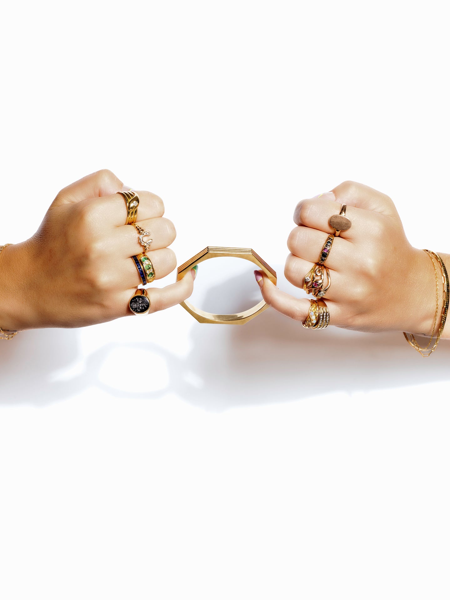 on a stark white background, two hands covered in gold rings appear to play tug of war with a geometric gold bangle