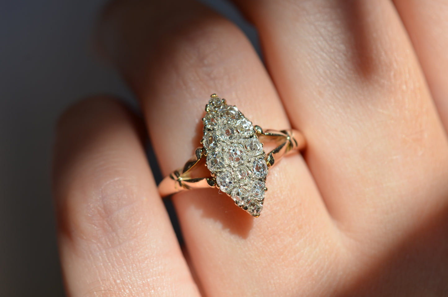 A closely cropped macro photo of the antique ring on a Caucasian model's left ring finger to show scale, photographed in natural light that dazzles on the diamonds, with the ring centered between two bands of shadows.