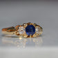 A Victorian ring in yellow gold with swirling shoulders and a central vivid blue sapphire flanked by a pair of old mine cut diamonds, photographed slightly to the right to focus on the details of the diamond facets.
