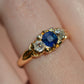 A closely cropped photo of the Victorian trilogy ring with an old mine cut diamond on either side of a vivid blue sapphire, all held in scrolling details of yellow gold. Shown on a Caucasian finger.