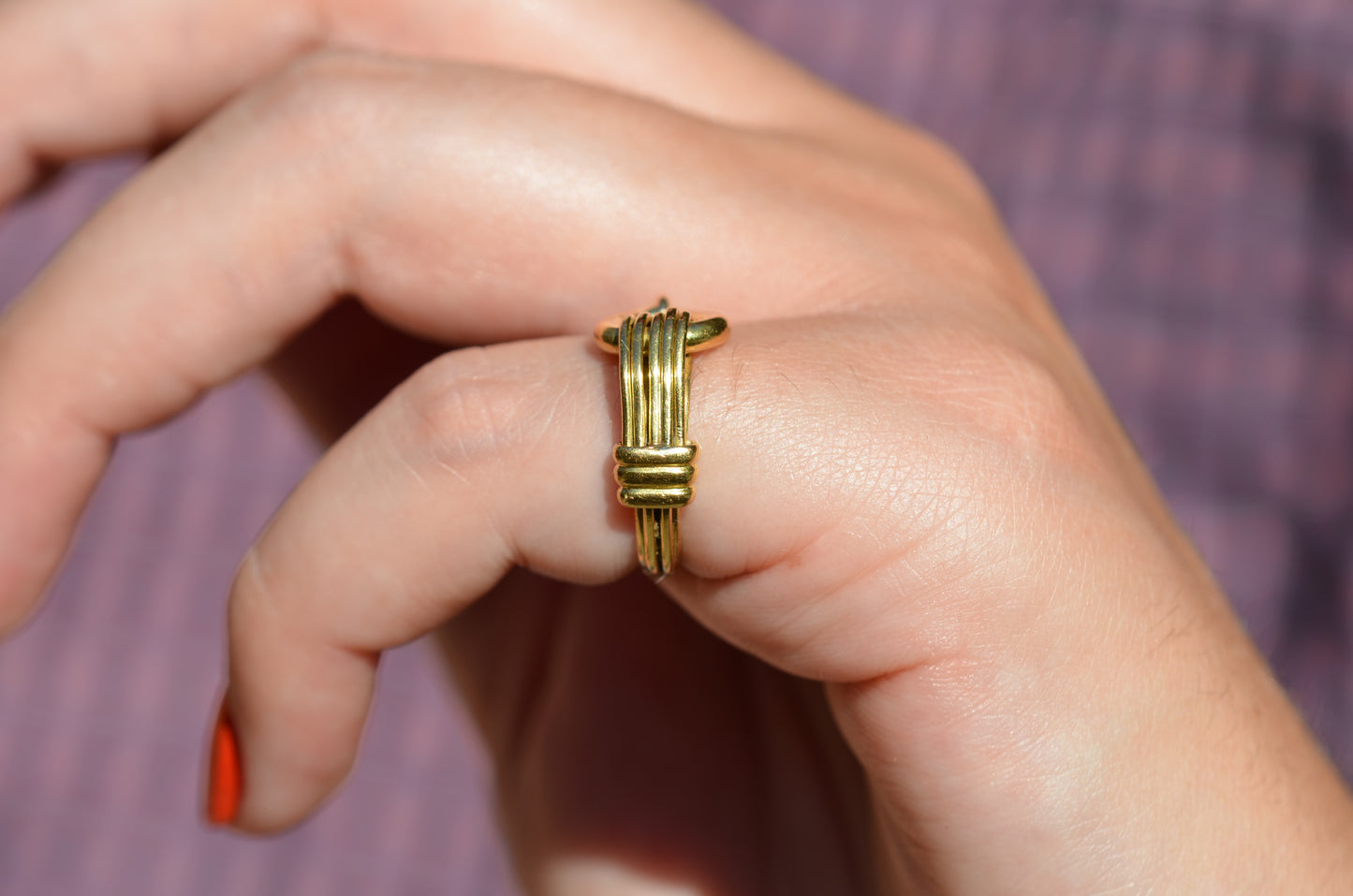 A medium-close view of the ring on a Caucasian model's left pinky finger. Her nail polish is an orangey-red, and in the background her blue and red gingham blouse is visible.