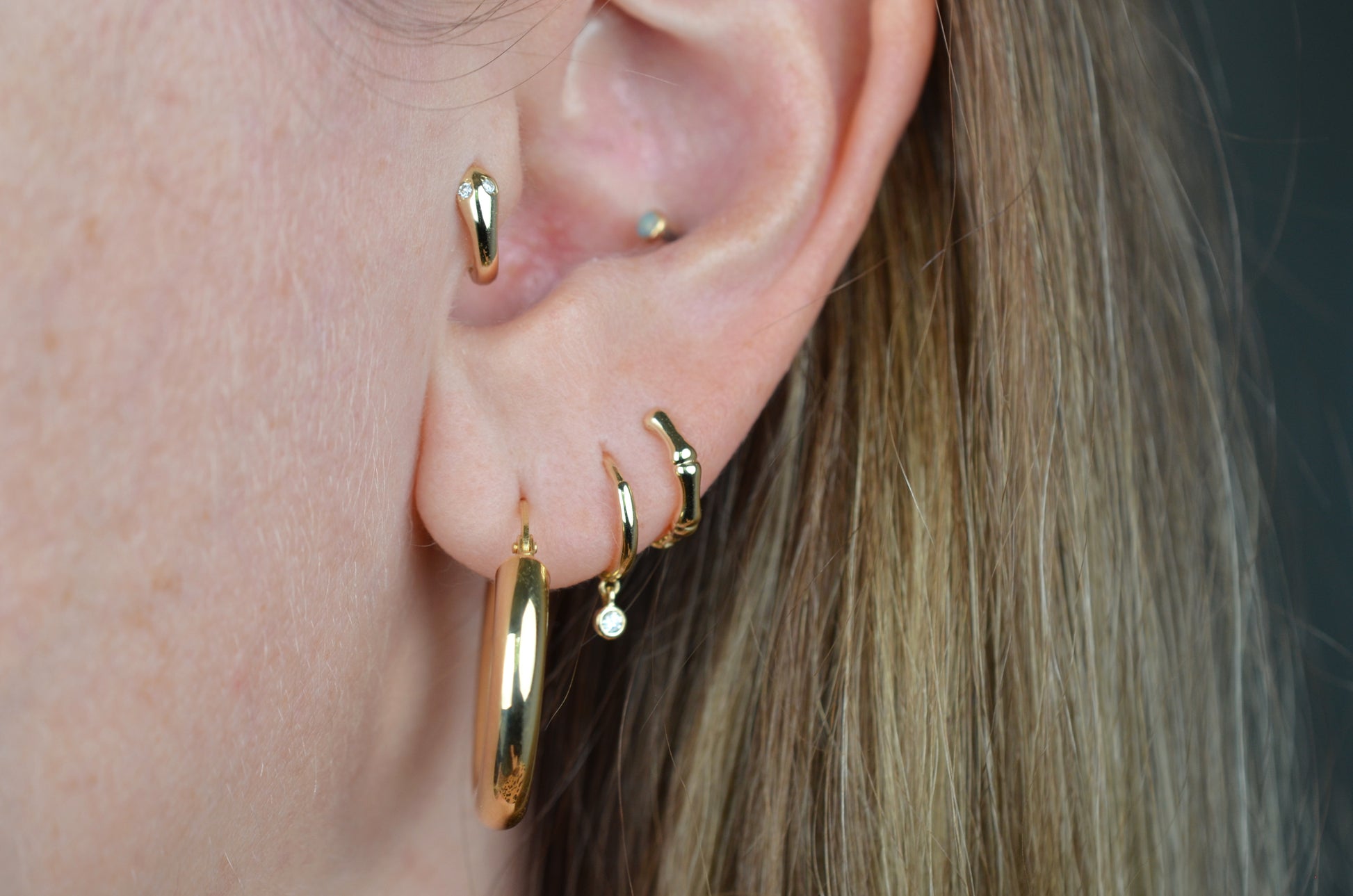 the featured hoops are shown on a caucasian model's first lobe piercing. also visible are a snug gold hoop with dangling diamond in the second piercing, a snug gold hoop stylized as bamboo in the third piercing, and a snug gold hoop stylized as a snake with diamond eyes in the tragus piercing.