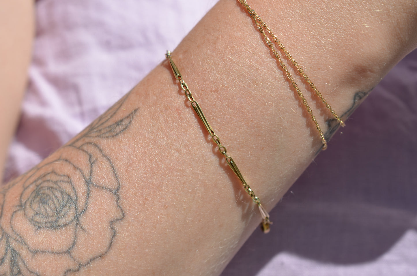 A closely cropped view of the bracelet on a Caucasian model's wrist. She is wearing several other gold bracelets and has a floral tattoo on her forearm.