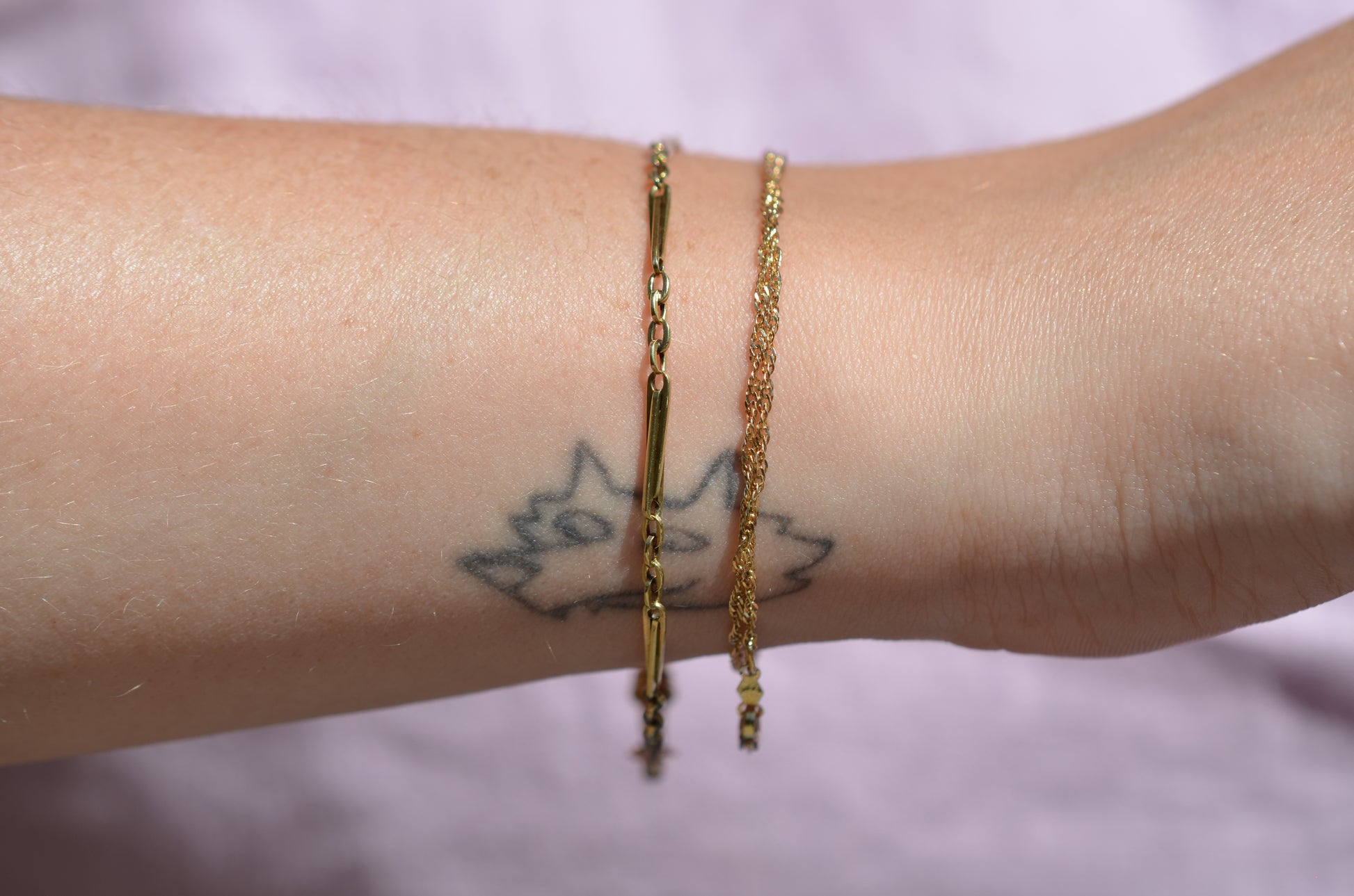 A closely cropped view of the bracelet on a Caucasian model's wrist. She is wearing several other gold bracelets and has a small tattoo of the "Space Coyote" character from The Simpsons.