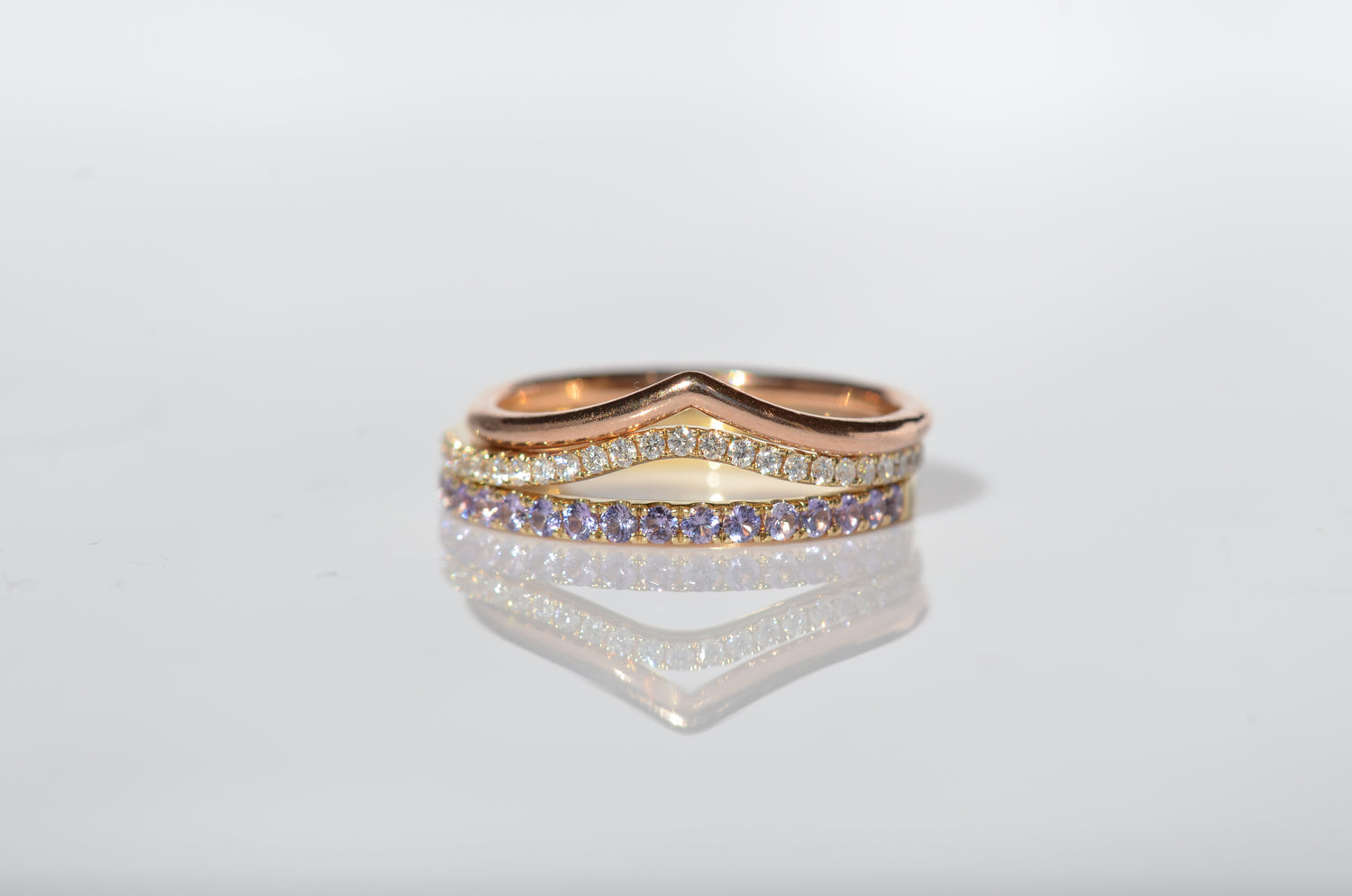 a minimal stack of three contemporary rings: at bottom, a pale purple sapphire eternity, above it a diamond eternity with contour, and on top a rose gold chevron band