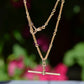 The elongated bar link Albert chain is suspended in front of a blurry dark green background dotted with pink florals. The t-bar of the necklace hangs in the centre, with the dog clip and bolt ring clasps visible.