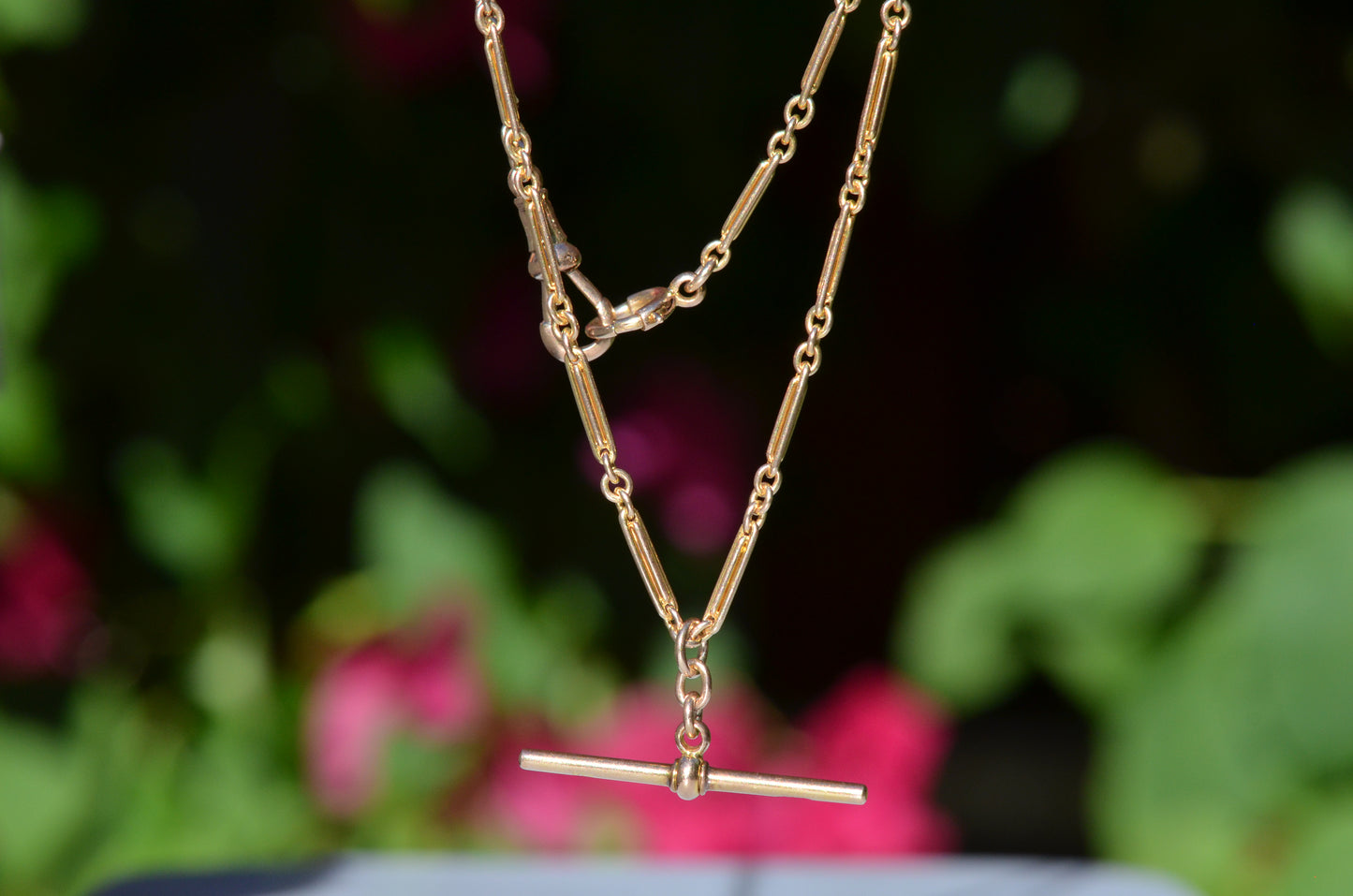 The elongated bar link Albert chain is suspended in front of a blurry dark green background dotted with pink florals. The t-bar of the necklace hangs in the centre, with the dog clip and bolt ring clasps visible.