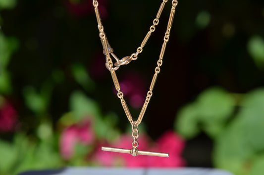 The elongated bar link Albert chain is suspended in front of a blurry dark green background dotted with pink florals. The t-bar of the necklace hangs in the centre, with the dog clip and bolt ring clasps visible.