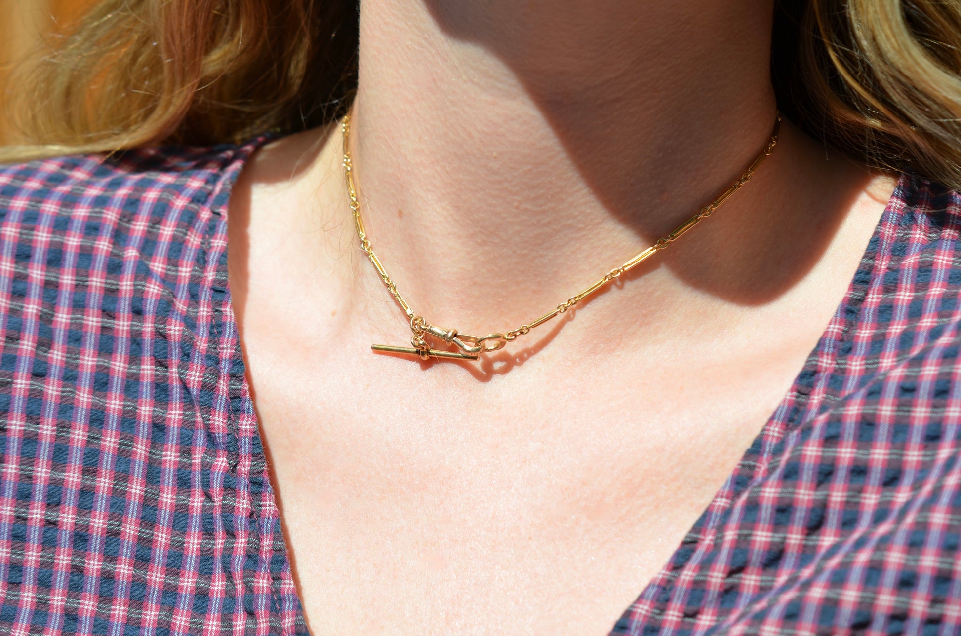 A Caucasian model wears the chain as a necklace, where it fits snugly at the base of her neck in a choker length. She is wearing a v-neck blue and red gingham blouse.