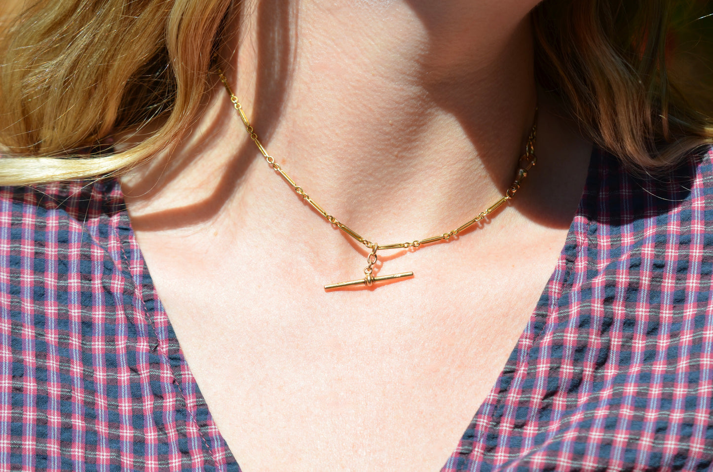 A Caucasian model wears the chain as a necklace, where it fits snugly at the base of her neck in a choker length. She is wearing a v-neck blue and red gingham blouse.