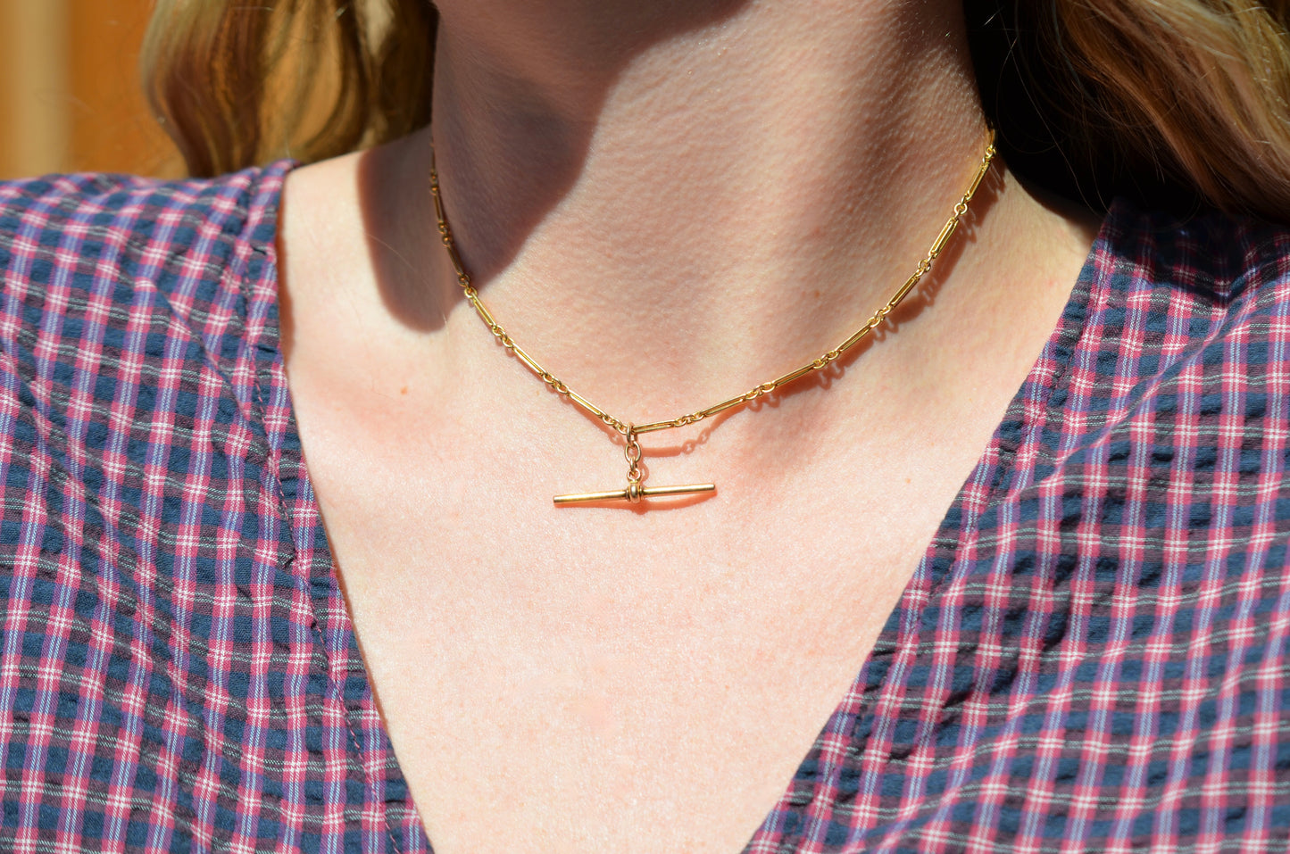 A Caucasian model wears the chain as a necklace, where it fits snugly at the base of her neck in a choker length. She is wearing a v-neck blue and red gingham blouse.