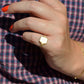 A medium-close view of the ring on a Caucasian model's left pinky finger. The model has bright orangey-red short nails and is wearing a red and blue gingham blouse.