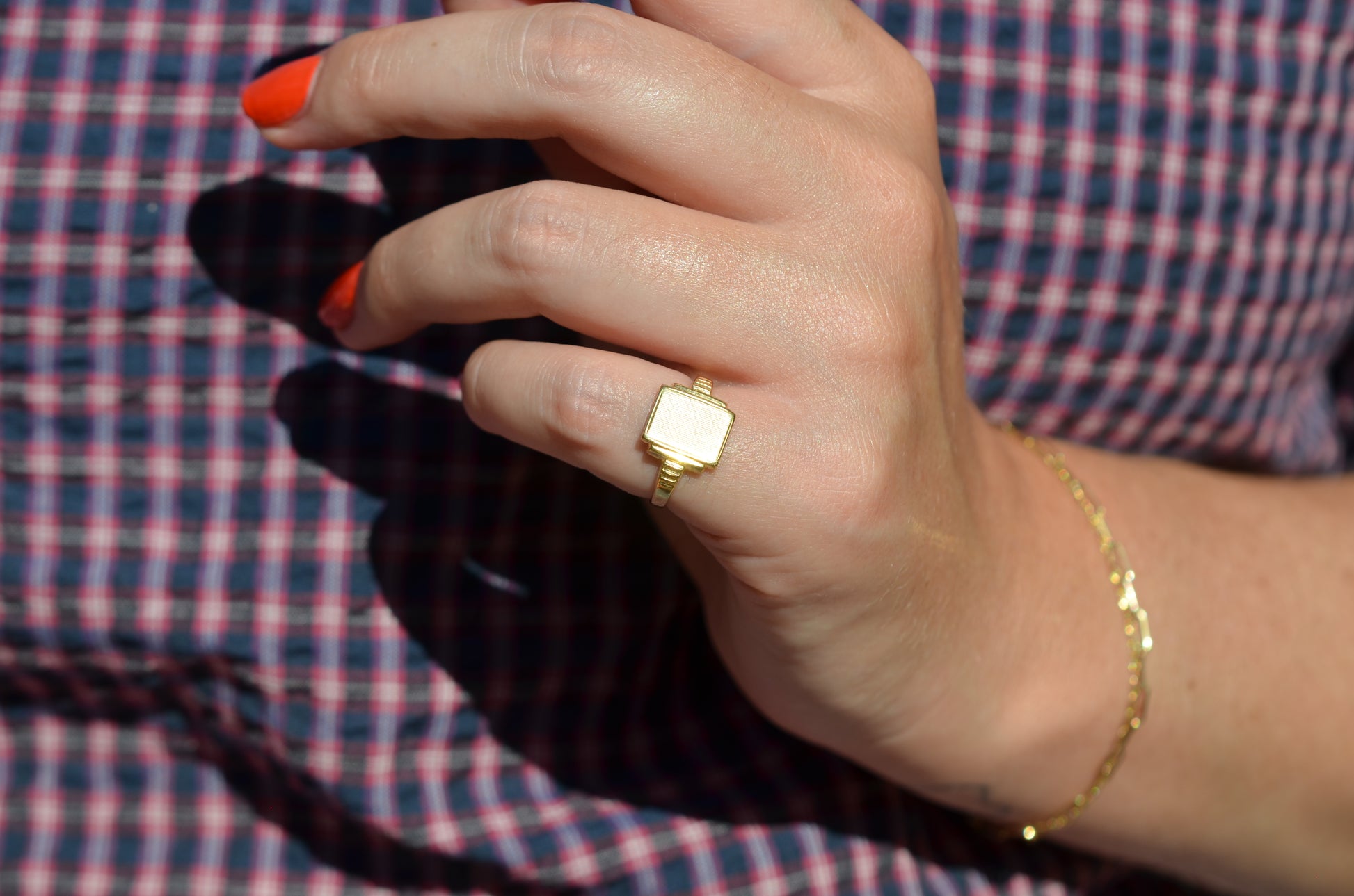 A medium-close view of the ring on a Caucasian model's left pinky finger. The model has bright orangey-red short nails and is wearing a red and blue gingham blouse.