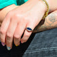 A medium-close view of the ring, vibrant in bright daylight, on the left pinky finger of a Caucasian model. The model has bright multicolored nails, a teal shirt and black jeans, and a floral tattoo visible on her arm.