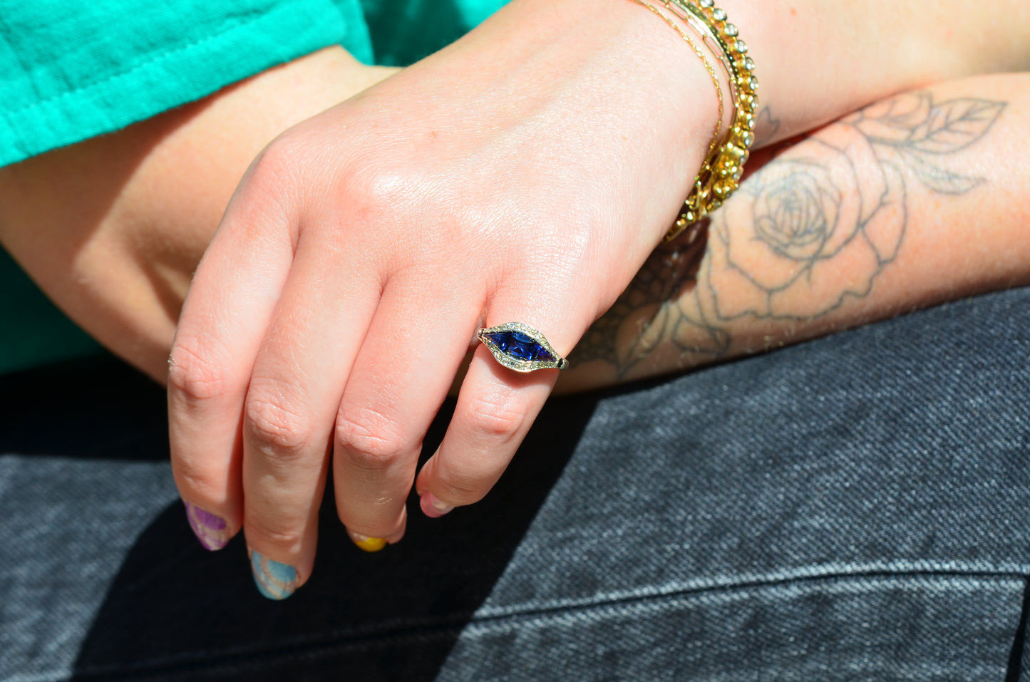 A medium-close view of the ring, vibrant in bright daylight, on the left pinky finger of a Caucasian model. The model has bright multicolored nails, a teal shirt and black jeans, and a floral tattoo visible on her arm.