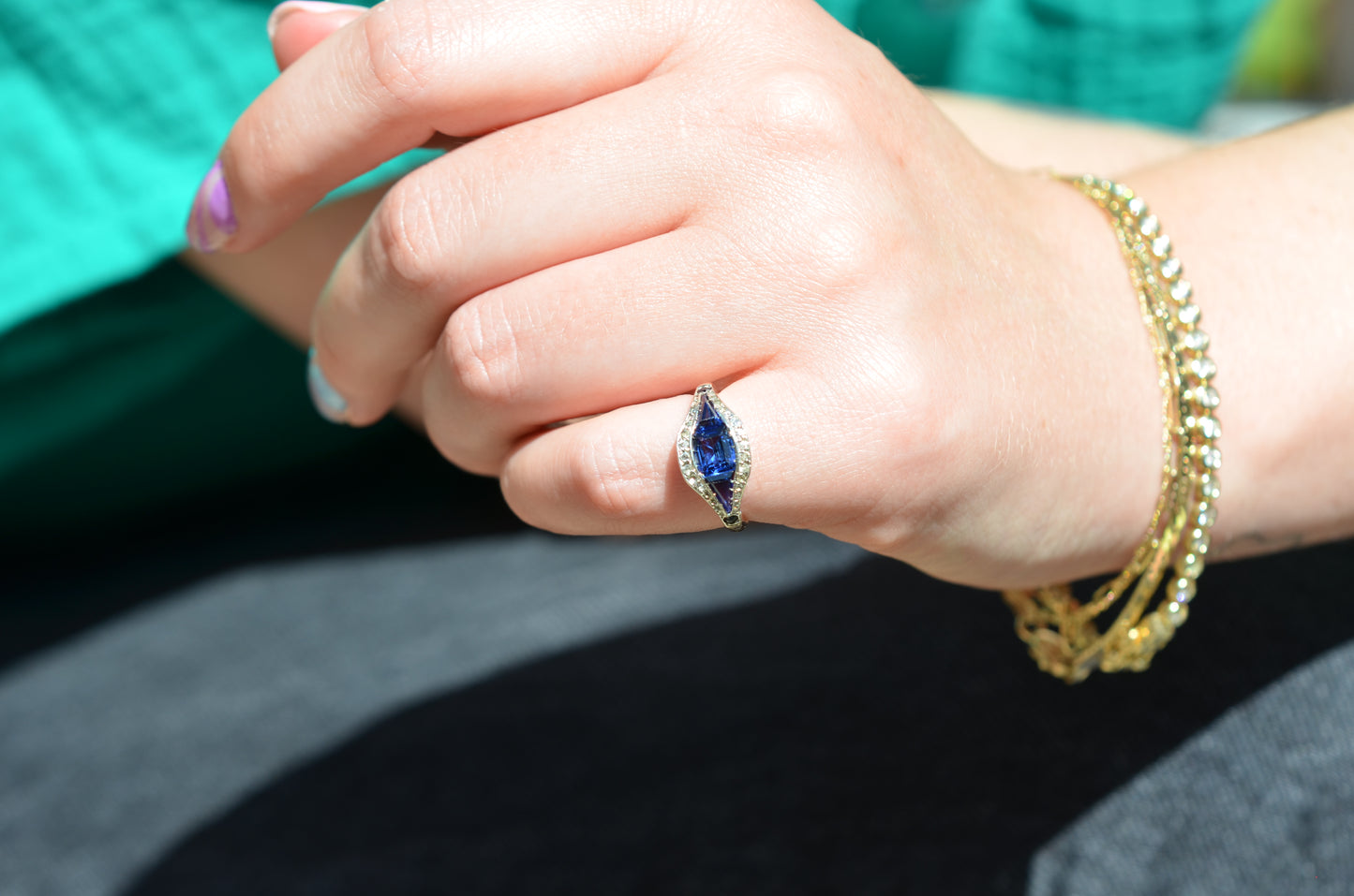 A medium-close view of the ring, vibrant in bright daylight, on the left pinky finger of a Caucasian model. The model has bright multicolored nails, a teal shirt and black jeans, and several gold bracelets.