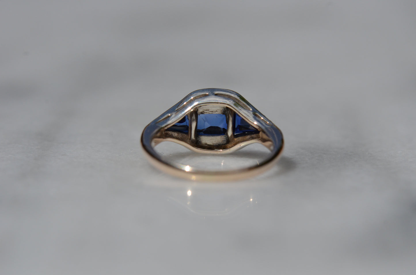 The ring, shown from behind and with the back of the head in focus, shows the open setting behind the blue past and solid, closed silver settings behind the colorless paste.