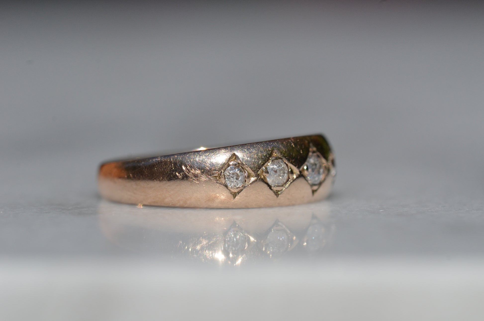 A tightly-cropped macro shot of the Victorian ring, showing the hand-cut diamond-shaped frames that hold each cushion-shaped stone. The old mine cut diamonds have internal fractures and inclusions but still sparkle in the direct sun, and the gold is weathered from a century of wear. The ring is facing slightly to the right, to better see the detail of the tapering side stones.