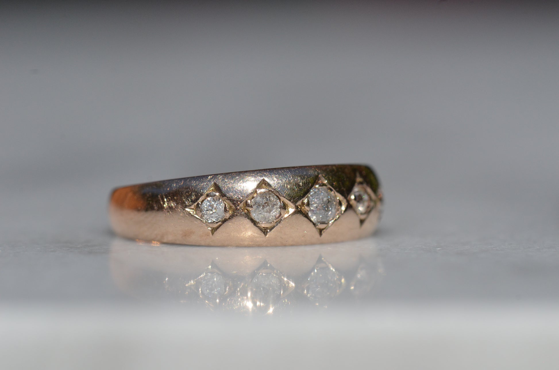 A tightly-cropped macro shot of the Victorian ring, showing the hand-cut diamond-shaped frames that hold each cushion-shaped stone. The old mine cut diamonds have internal fractures and inclusions but still sparkle in the direct sun, and the gold is weathered from a century of wear. The ring is facing slightly to the right, to better see the detail of the tapering side stones.