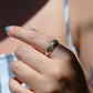 A medium-close view of the ring on the left pointer finger of a Caucasian model. Her hand rests in the foreground, while in the background she wears a striped top with thin straps. The edge of a floral tattoo is visible on her upper arm.