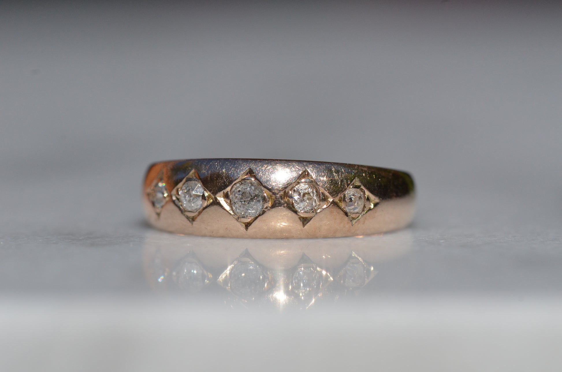 A tightly-cropped macro shot of the Victorian ring, showing the hand-cut diamond-shaped frames that hold each cushion-shaped stone. The old mine cut diamonds have internal fractures and inclusions but still sparkle in the direct sun, and the gold is weathered from a century of wear. The ring is facing slightly to the left, to better see the detail of the tapering side stones.