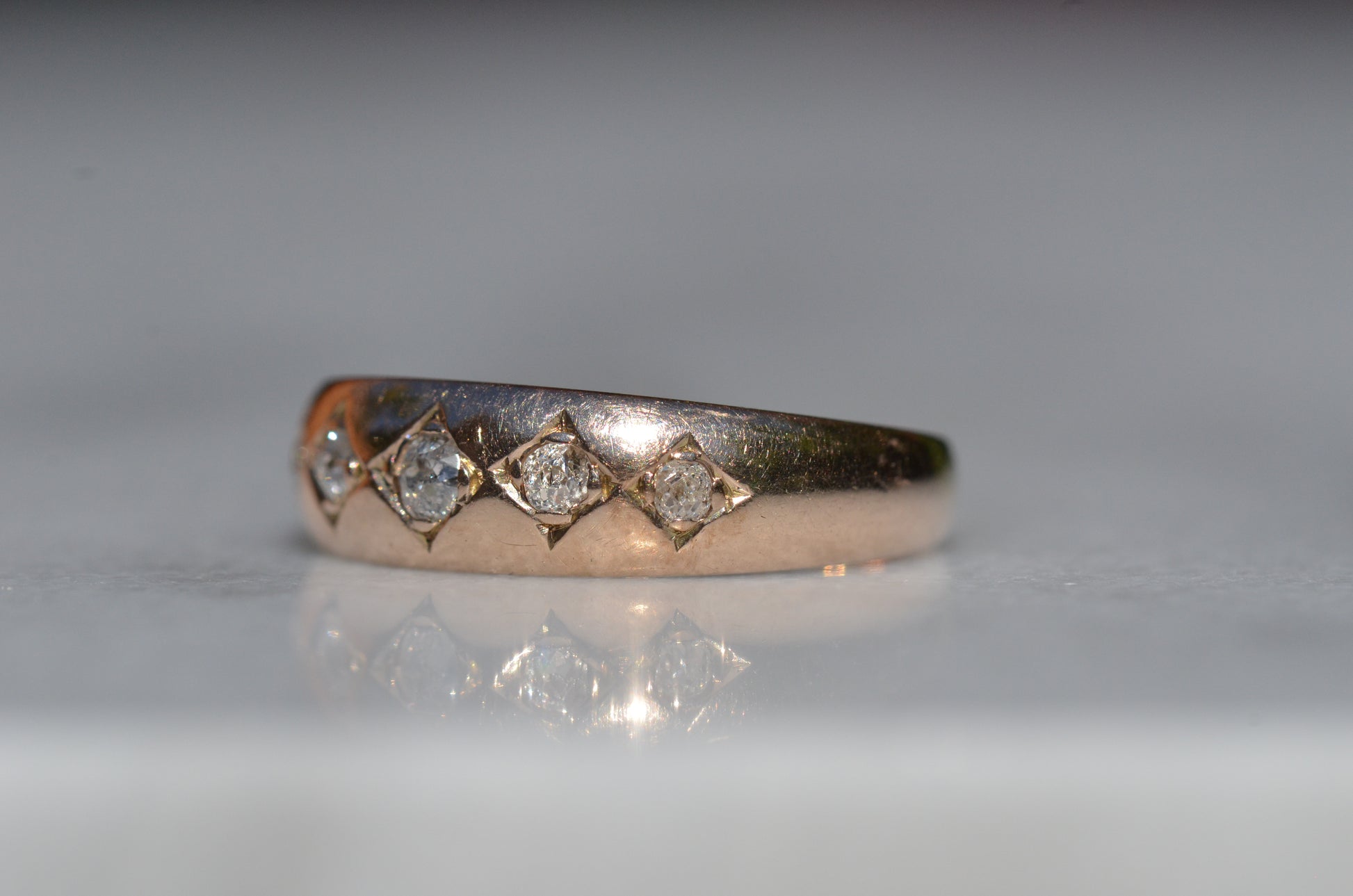 A tightly-cropped macro shot of the Victorian ring, showing the hand-cut diamond-shaped frames that hold each cushion-shaped stone. The old mine cut diamonds have internal fractures and inclusions but still sparkle in the direct sun, and the gold is weathered from a century of wear. The ring is facing slightly to the left, to better see the detail of the tapering side stones.