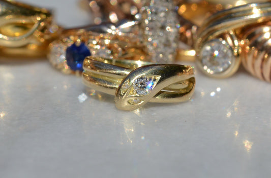 Macro of a Victorian yellow gold snake ring in front of an out-of-focus field of antique gold rings. The direct sun has caused a flash of white, blue, and violet to dazzle from the central old European cut diamond in the snake's head.