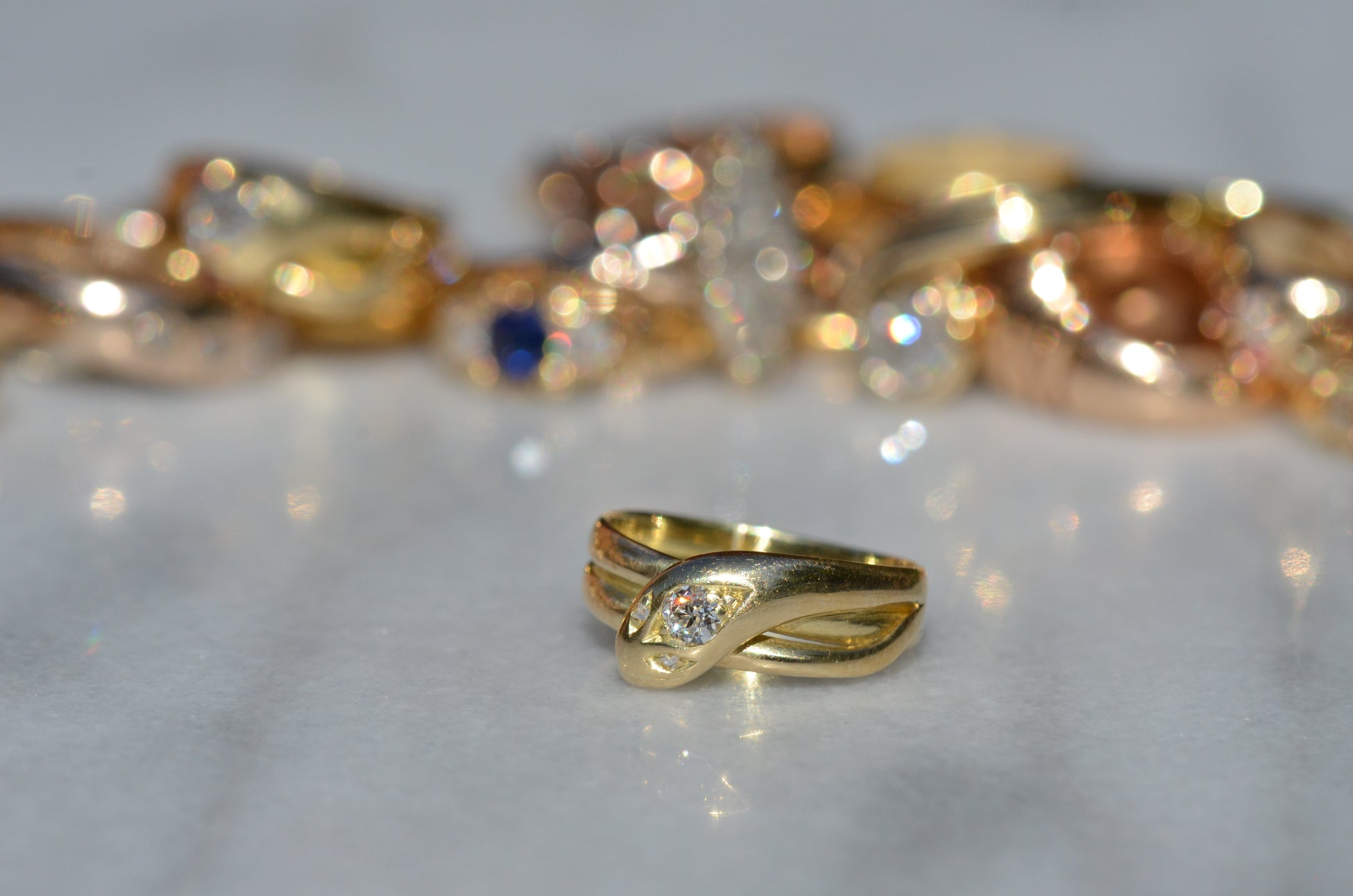 Macro of a Victorian yellow gold snake ring in front of an out-of-focus field of antique gold rings. The direct sun has caused a flash of white, orange, and red to dazzle from the central old European cut diamond in the snake's head.