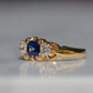A Victorian ring in yellow gold with swirling shoulders and a central vivid blue sapphire flanked by a pair of old mine cut diamonds, photographed turned slightly to the left.