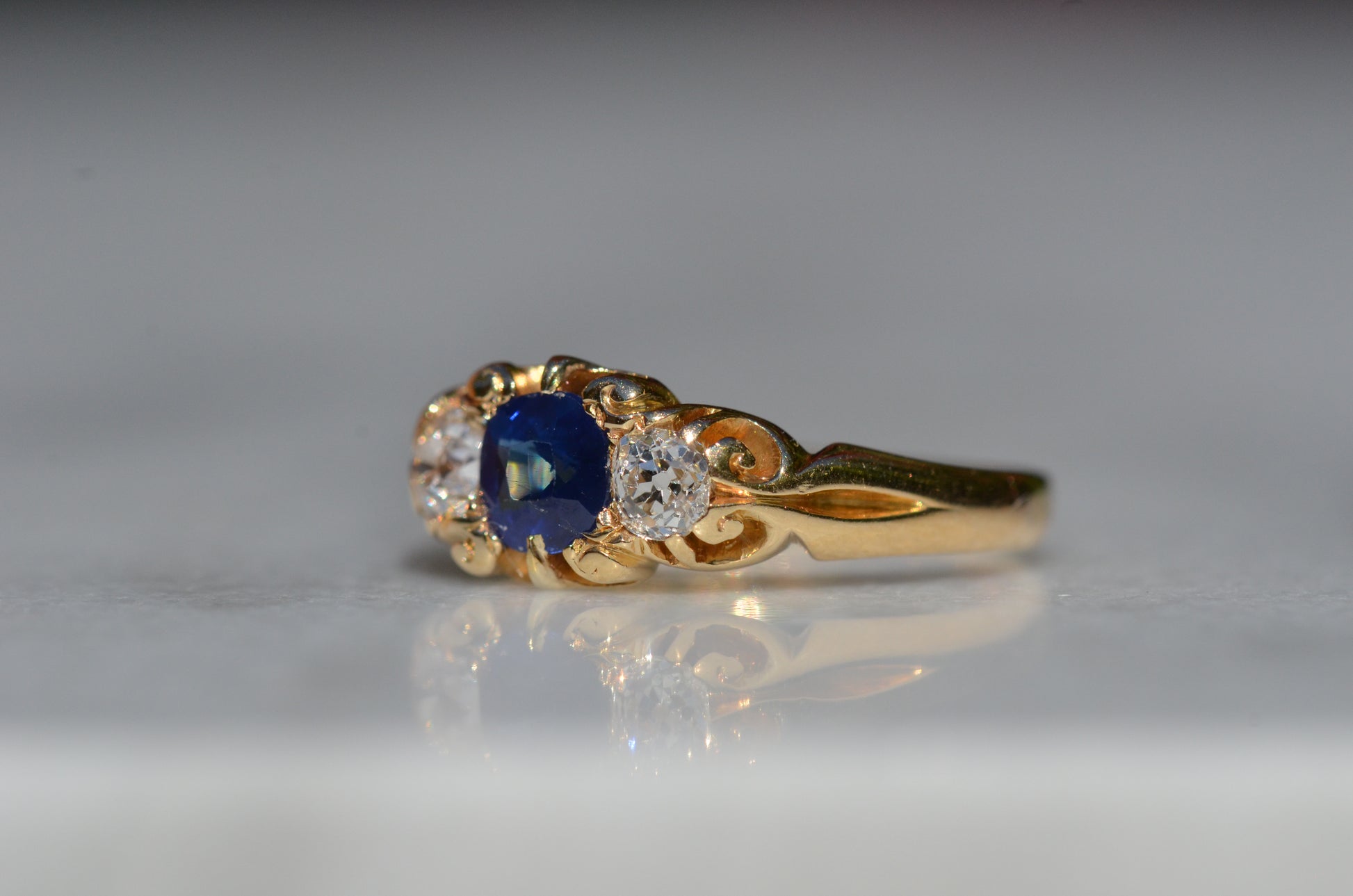 A Victorian ring in yellow gold with swirling shoulders and a central vivid blue sapphire flanked by a pair of old mine cut diamonds, photographed turned slightly to the left.