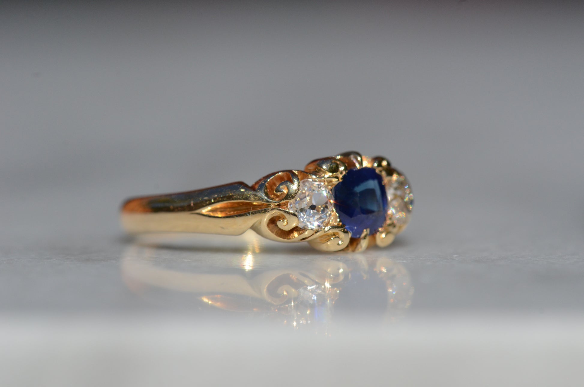 A Victorian ring in yellow gold with swirling shoulders and a central vivid blue sapphire flanked by a pair of old mine cut diamonds, photographed turned to the right to highlight the side view of the ring.