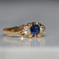 A Victorian ring in yellow gold with swirling shoulders and a central vivid blue sapphire flanked by a pair of old mine cut diamonds, photographed moderately to the right to highlight to shoulder of the ring.