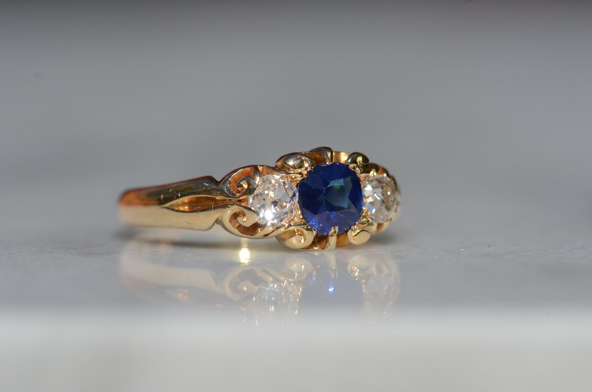 A Victorian ring in yellow gold with swirling shoulders and a central vivid blue sapphire flanked by a pair of old mine cut diamonds, photographed moderately to the right to highlight to shoulder of the ring.