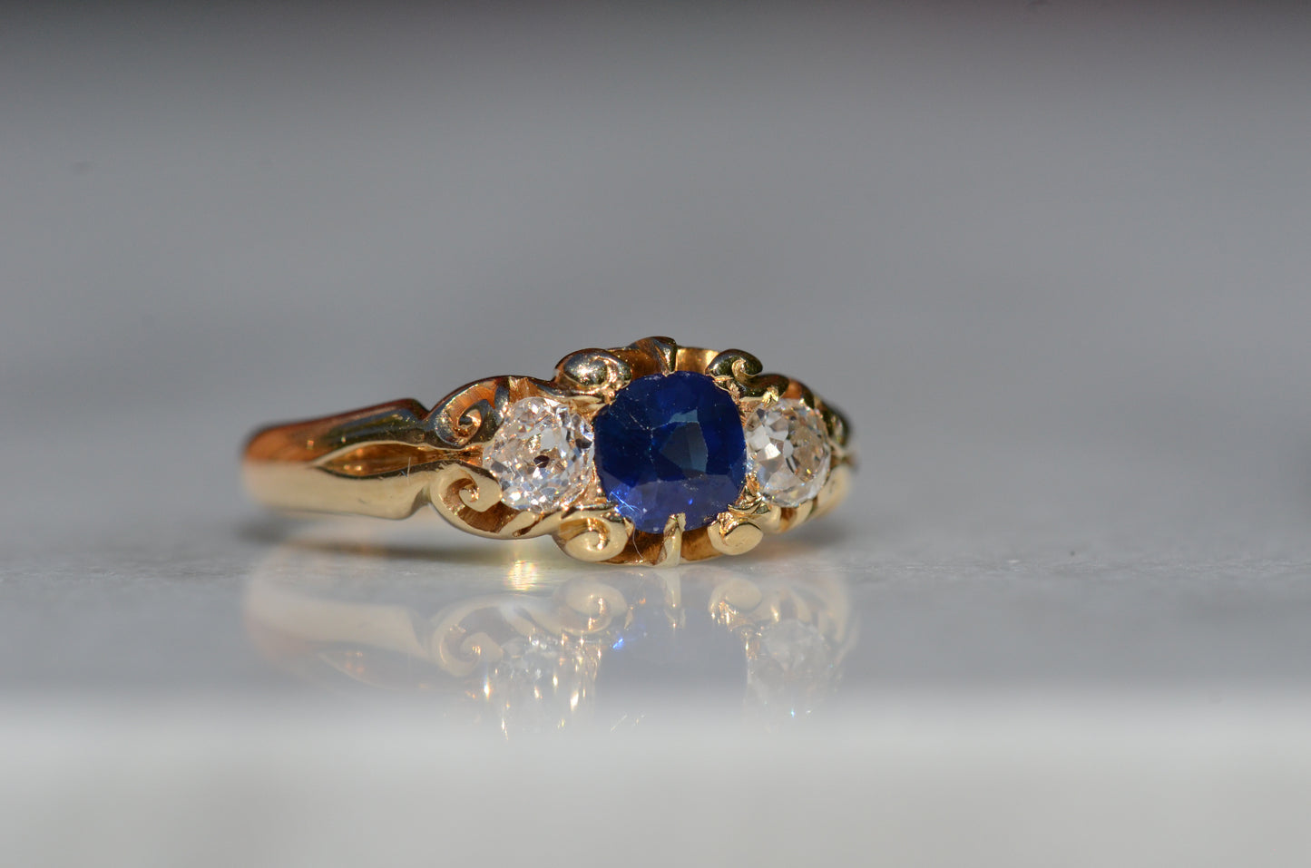 A Victorian ring in yellow gold with swirling shoulders and a central vivid blue sapphire flanked by a pair of old mine cut diamonds, photographed slightly to the right to focus on the details of the diamond facets.