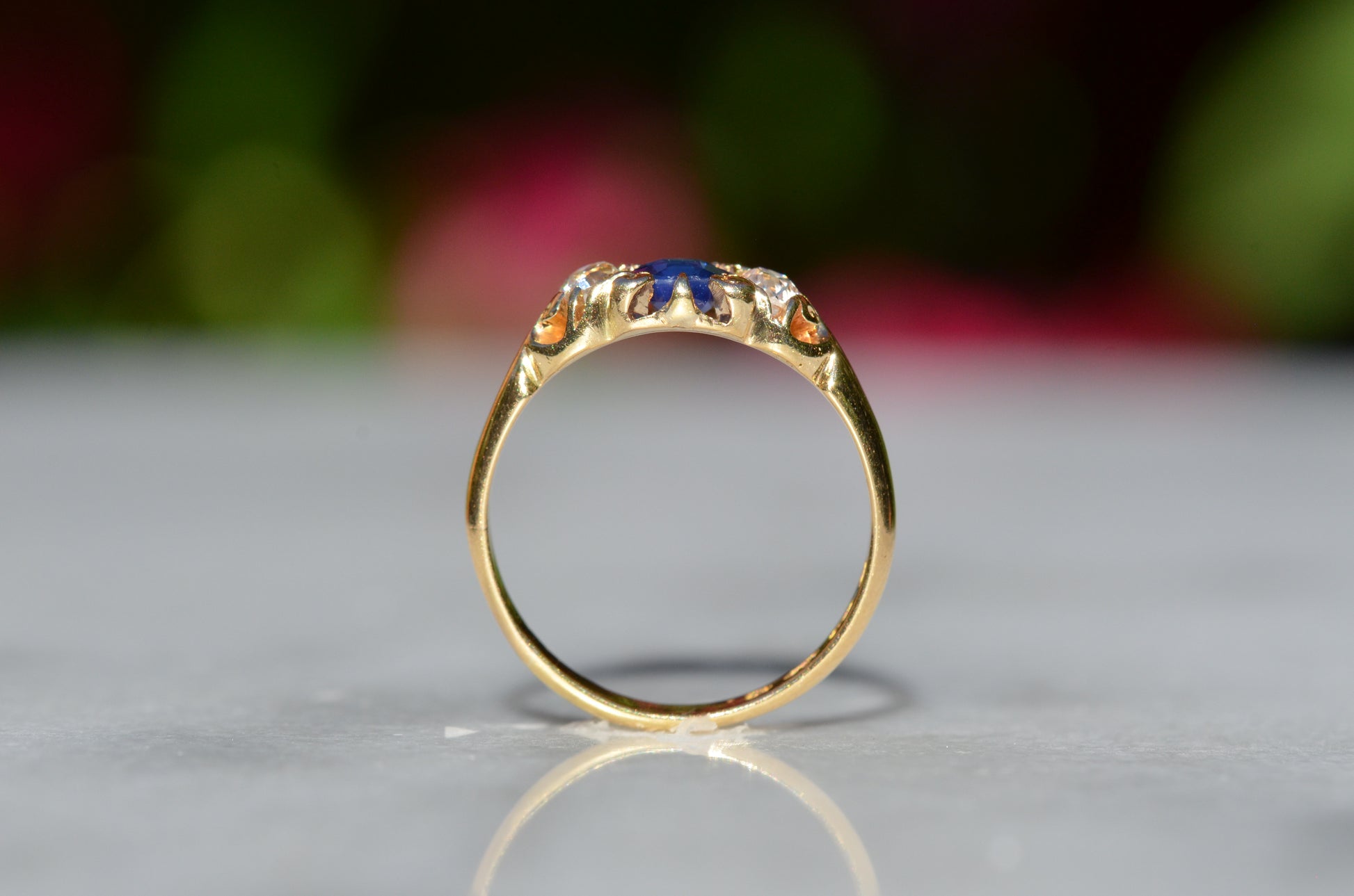 A Victorian ring in yellow gold with swirling shoulders and a central vivid blue sapphire flanked by a pair of old mine cut diamonds, photographed positioned upright looking through the finger hole to showcase the gallery detail and rise off the finger.