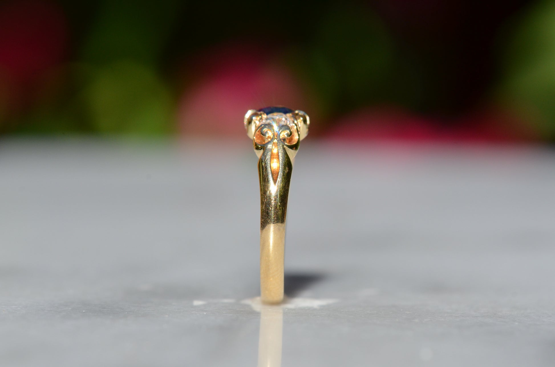 A Victorian ring in yellow gold with swirling shoulders and a central vivid blue sapphire flanked by a pair of old mine cut diamonds, photographed upright and from the side, to showcase the profile of the ring.