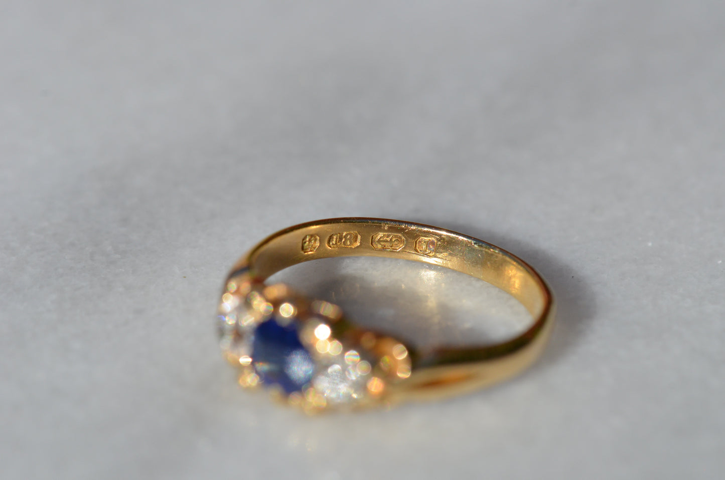 A Victorian ring in yellow gold with swirling shoulders and a central vivid blue sapphire flanked by a pair of old mine cut diamonds, photographed looking into the inside of the band to showcase the partial set of British hallmark stamps.