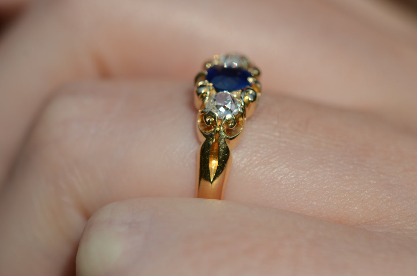 A closely cropped photo of the Victorian trilogy ring with an old mine cut diamond on either side of a vivid blue sapphire, all held in scrolling details of yellow gold. Shown on a Caucasian finger in a side view to highlight the ring's shoulder.