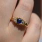 A closely cropped photo of the Victorian trilogy ring with an old mine cut diamond on either side of a vivid blue sapphire, all held in scrolling details of yellow gold. Shown on a Caucasian finger.