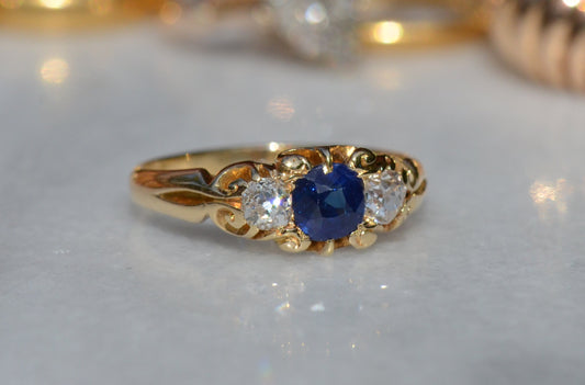 A tightly cropped macro photo showcases the rainbow dispersion from an old mine cut diamond, a pair of which flank a velvety blue sapphire, all held in a scrolling yellow gold ring.