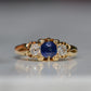 A Victorian ring in yellow gold with swirling shoulders and a central vivid blue sapphire flanked by a pair of old mine cut diamonds, photographed head-on.