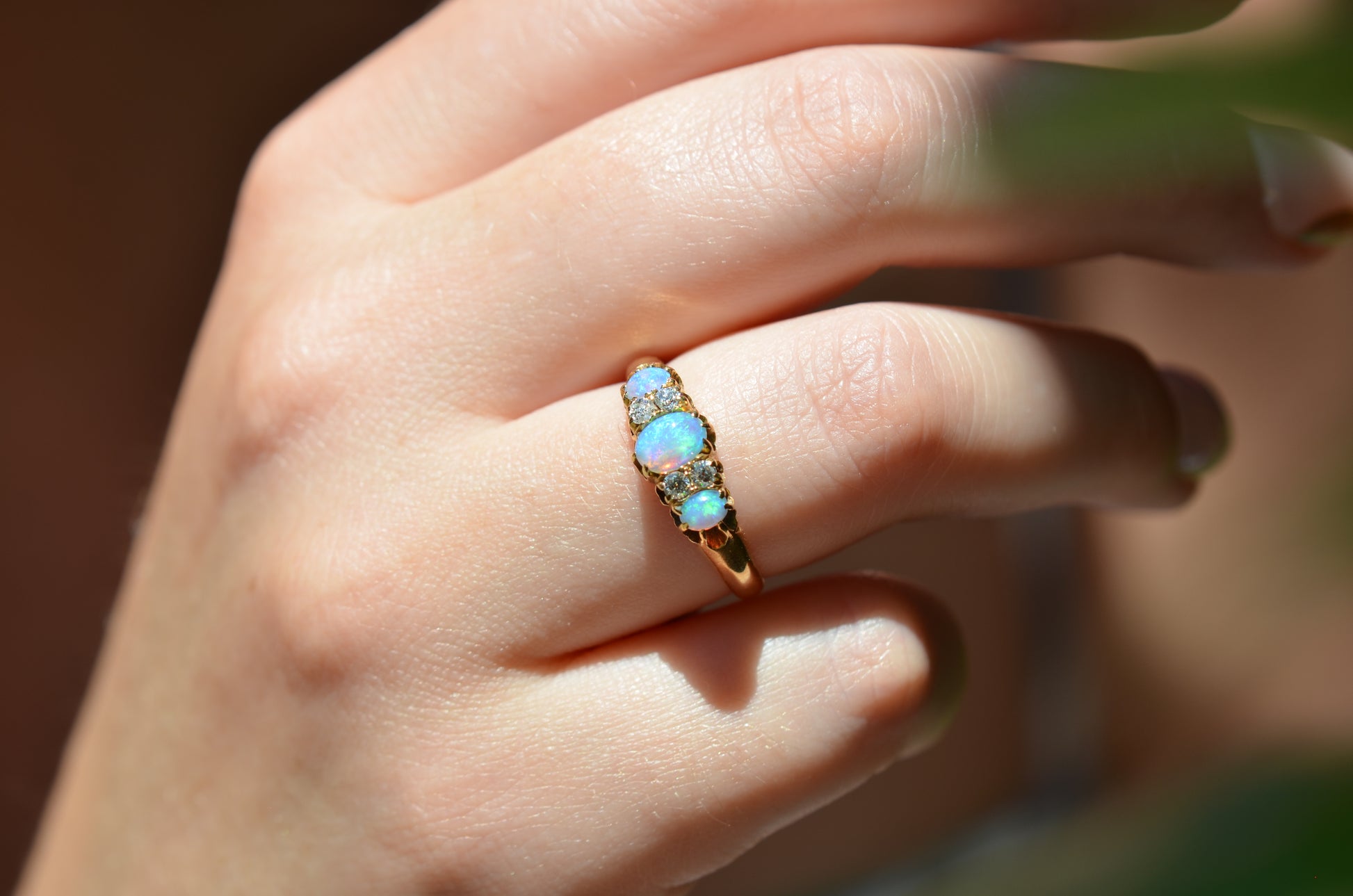 A very tightly cropped macro of the ring on a Caucasian model, showing the scale to the ring on the hand as well as the play of color of the opals in direct sunlight. While the most prominent colors are blue with green and violet, a flame of orange is visible to one side.