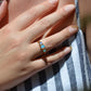 A medium-close image of the ring on a Caucasian model, showing the scale to the ring on the hand as well as the play of color of the opals in direct sunlight. While the most prominent colors are blue with green and violet, a flame of orange is visible to one side. The model's hand is resting on her own chest; also visible are long pink and gold nails and the edge of a floral arm tattoo.