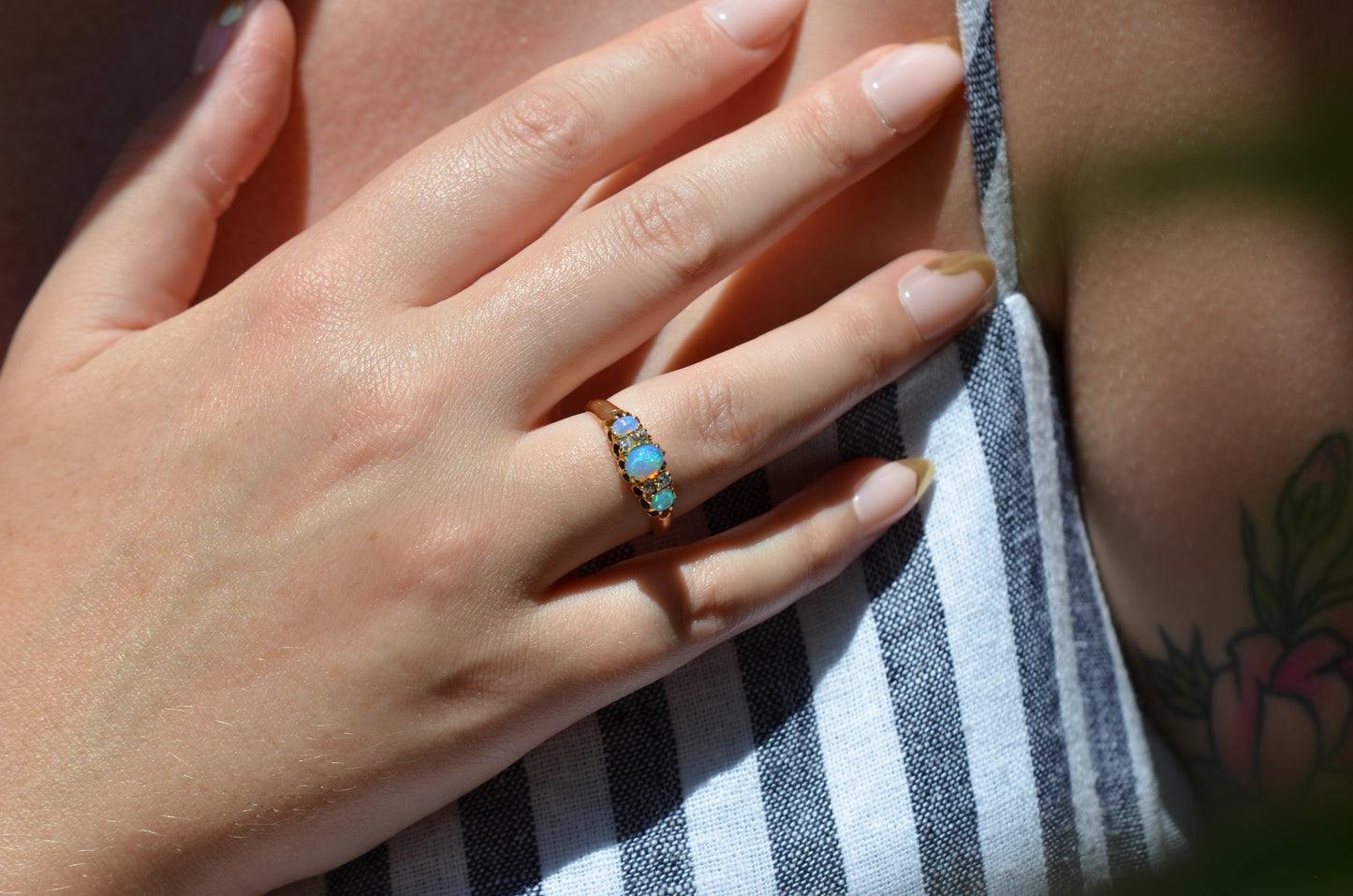 A medium-close image of the ring on a Caucasian model, showing the scale to the ring on the hand as well as the play of color of the opals in direct sunlight. While the most prominent colors are blue with green and violet, a flame of orange is visible to one side. The model's hand is resting on her own chest; also visible are long pink and gold nails and the edge of a floral arm tattoo.