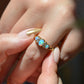 A very tightly cropped macro of the ring on a Caucasian model, showing the scale to the ring on the hand as well as the play of color of the opals in direct sunlight. While the most prominent colors are blue with green and violet, a flame of orange is visible to one side. The model's other hand, with long pink and gold fingernails, is easing the ring onto the finger.