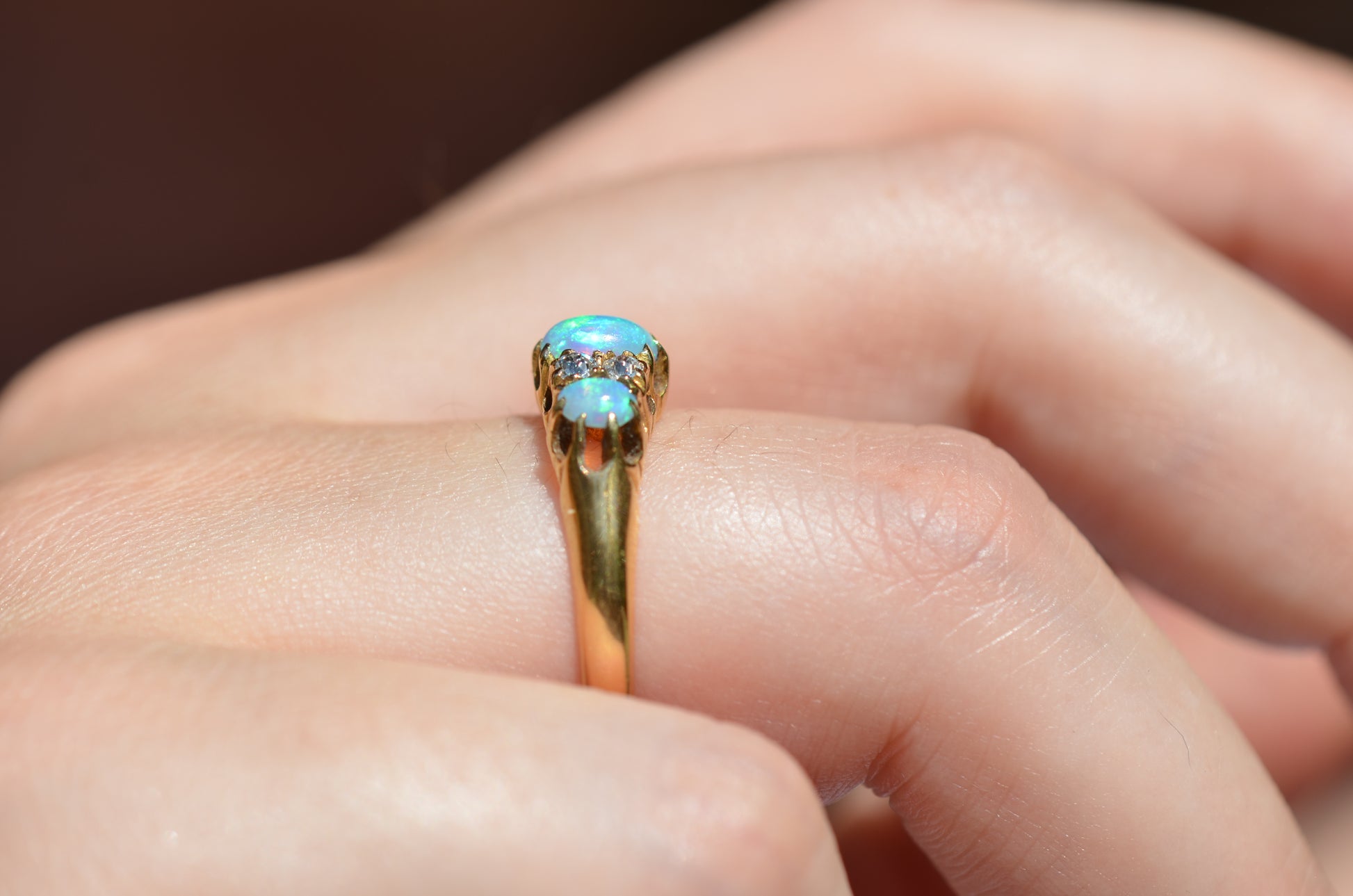 A very tightly cropped macro of the ring on a Caucasian model, showing the scale to the ring on the hand as well as the play of color of the opals in direct sunlight. While the most prominent colors are blue with green and violet, a flame of orange is visible to one side. The ring is photographed from the side to show the low rise off the finger.