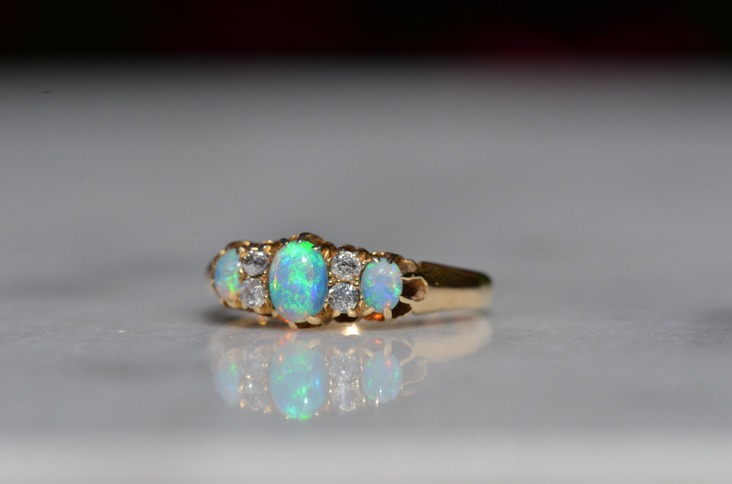 A closely-cropped macro of the Victorian ring, showcasing the strong green and blue play of color in the opals with a hint of violet and orange. The ring is photographed turned slightly to the left, so that the faceting and open culets of the old European diamonds are also visible.