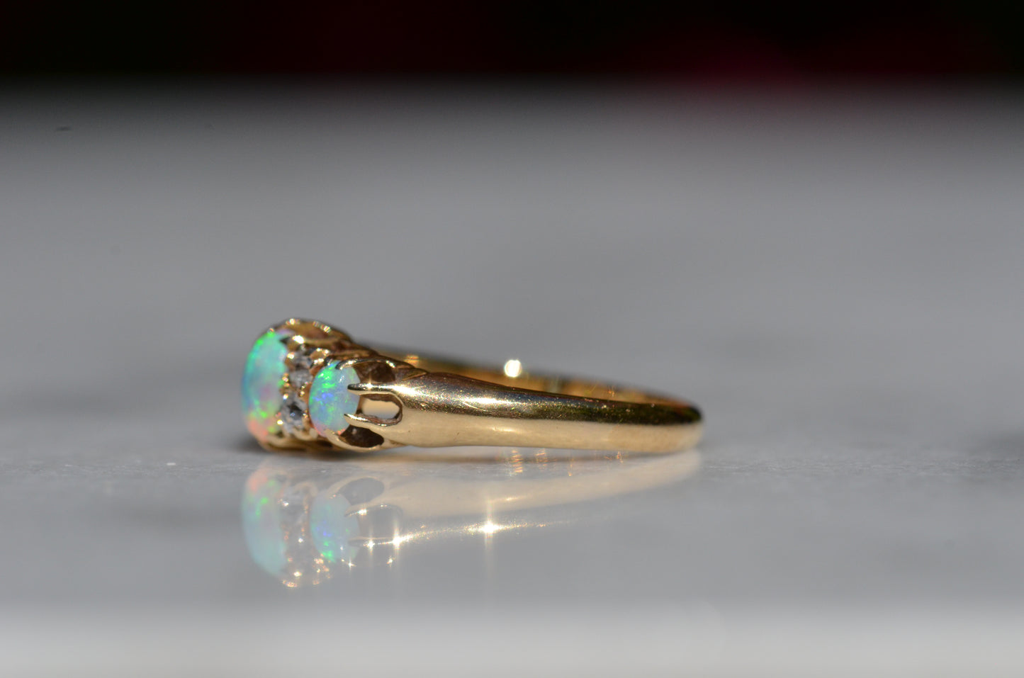 A closely-cropped macro of the Victorian ring, showcasing the strong green and blue play of color in the opals with a hint of violet and orange. The ring is photographed turned to the left to better showcase the slim prongs and buttery gold band.