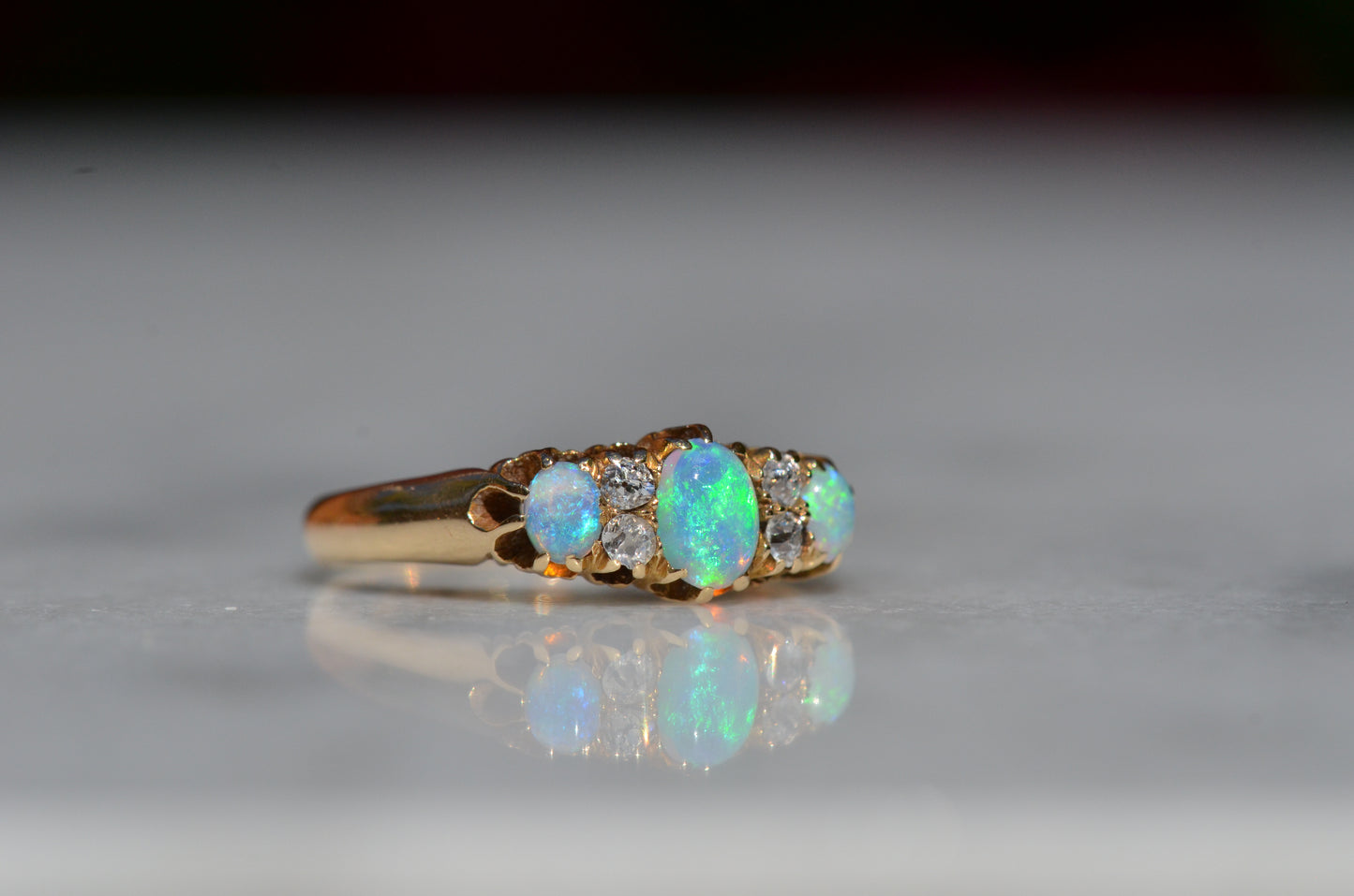 A closely-cropped macro of the Victorian ring, showcasing the strong green and blue play of color in the opals with a hint of violet and orange. The ring is photographed turned slightly to the right, so that the unique facet patterns of the accent diamonds are highlighted.