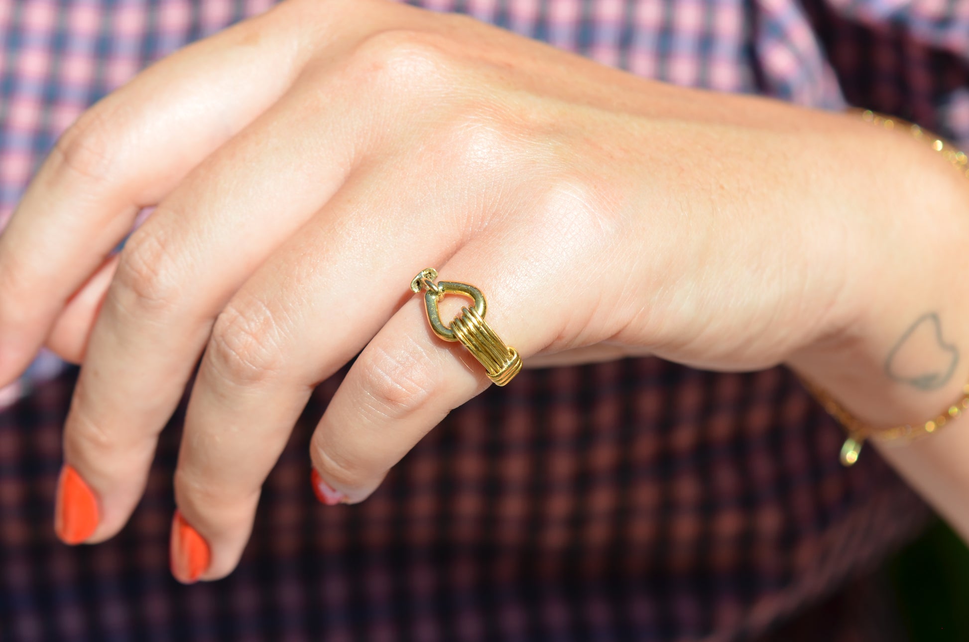 A medium-close view of the ring on a Caucasian model's left pinky finger. Her nail polish is an orangey-red, and in the background her blue and red gingham blouse is visible.